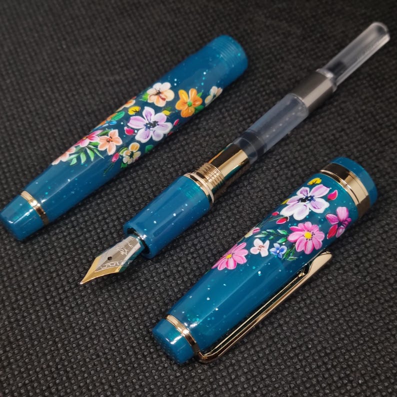 May include: A teal fountain pen featuring a floral pattern. The pen's body and cap are decorated with hand-painted flowers in pink, orange, and white. Gold-coloured trim and a gold nib are also visible. The pen is shown in pieces.