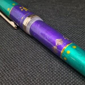 K&ouml;nnte beinhalten: Nahaufnahme eines dekorativen Stifts mit einem t&uuml;rkis- und lilafarbenen Schaft. Der Stift hat goldfarbene Akzente, darunter ein Clip und dekorative Muster. Der Schaft hat einen metallischen Glanz, und das Design umfasst gelbe Punkte und geometrische Formen.