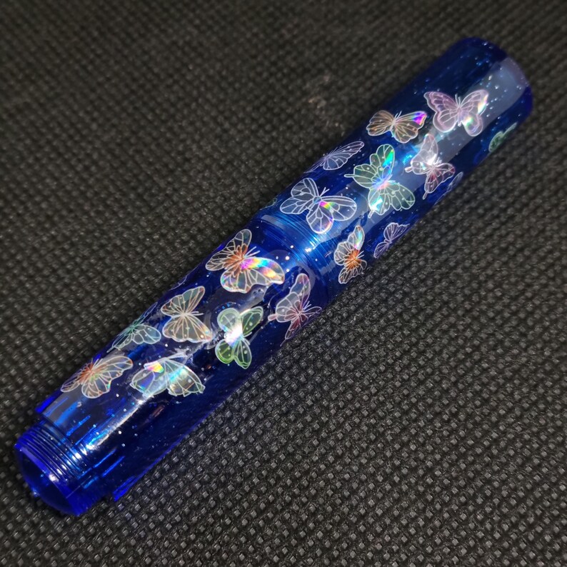 Puede incluir: Bol&iacute;grafo azul con dise&ntilde;o de mariposas iridiscentes. El bol&iacute;grafo es de color azul oscuro con un acabado brillante. Las mariposas parecen flotar sobre la superficie. El bol&iacute;grafo tiene un extremo roscado, mide unos 15 cm.