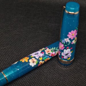 May include: A teal fountain pen with a floral design. The pen features a cap and barrel adorned with colorful flowers in pink, orange, white, and yellow. Gold-coloured accents are visible on the pen's trim.
