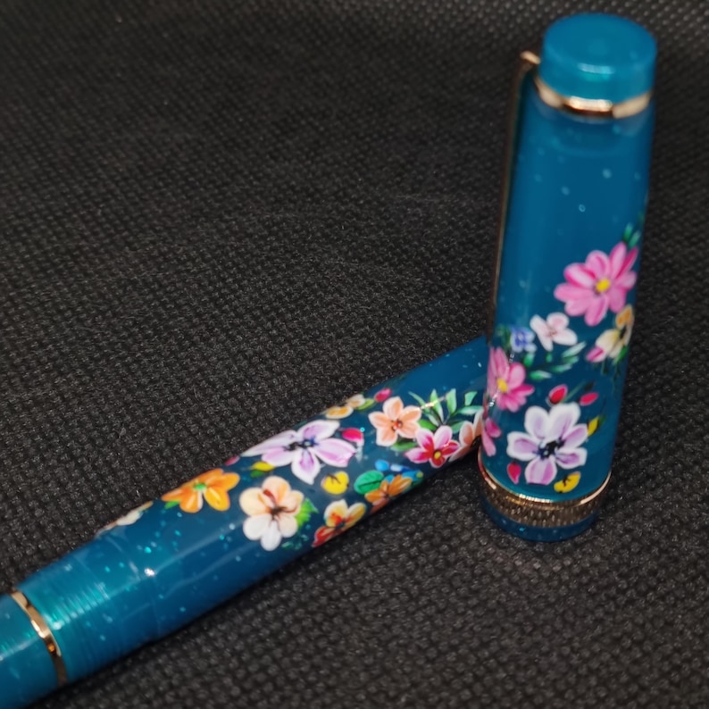 May include: A teal fountain pen with a floral design. The pen features a cap and barrel adorned with colorful flowers in pink, orange, white, and yellow. Gold-coloured accents are visible on the pen's trim.