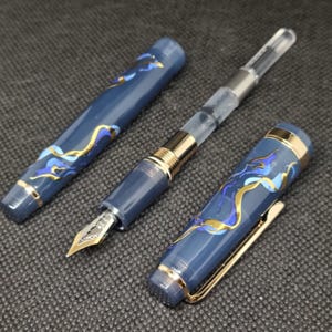 Könnte beinhalten: Dunkelblauer Füllfederhalter mit goldenen und blauen Zierakzenten. Der Stift hat eine goldene Feder und einen goldenen Clip. Das Design des Stifts zeigt fließende Linien in Gold und Blau auf dunklem Blau. Der Stift wird ohne Kappe und zerlegt gezeigt.