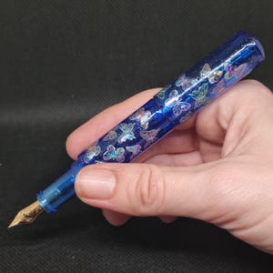 Puede incluir: Una pluma estilogr&aacute;fica azul con plum&iacute;n dorado y cuerpo azul transparente. El cuerpo est&aacute; decorado con calcoman&iacute;as de mariposas iridiscentes. El bol&iacute;grafo se sostiene en una mano sobre un fondo oscuro.
