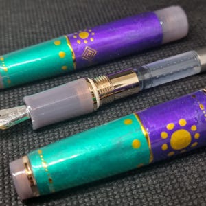 K&ouml;nnte beinhalten: Nahaufnahme von drei dekorativen F&uuml;llfederhaltern. Die Stifte haben t&uuml;rkisfarbene und violette Sch&auml;fte mit goldenen Akzenten und Mustern. Die Federn sind silberfarben und die Kappen durchscheinend grau.