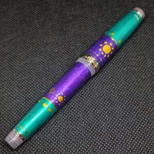 K&ouml;nnte beinhalten: Dekorativer F&uuml;llfederhalter mit einem t&uuml;rkis-violetten Schaft. Der Stift hat goldfarbene Akzente und ein Muster aus gelben Sonnen und geometrischen Formen. Der Stift hat eine silberfarbene Spitze und Endkappe.