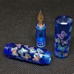 Puede incluir: Pluma estilogr&aacute;fica azul desmontada con plum&iacute;n dorado. El cuerpo y el capuch&oacute;n de la pluma est&aacute;n decorados con dise&ntilde;os de mariposas iridiscentes. La pluma est&aacute; hecha de un material azul transl&uacute;cido con purpurina.