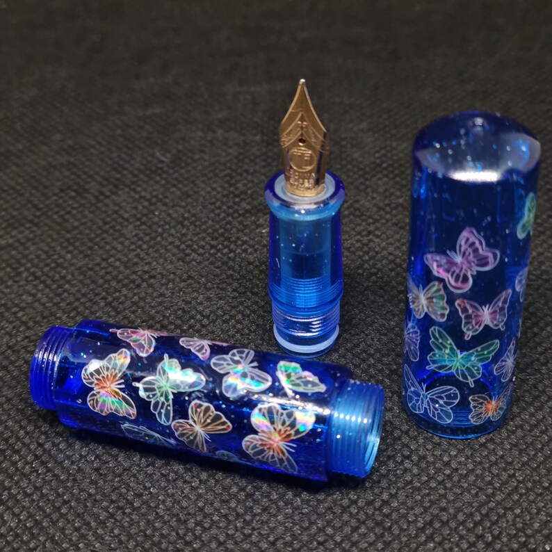 Puede incluir: Pluma estilogr&aacute;fica azul desmontada con plum&iacute;n dorado. El cuerpo y el capuch&oacute;n de la pluma est&aacute;n decorados con dise&ntilde;os de mariposas iridiscentes. La pluma est&aacute; hecha de un material azul transl&uacute;cido con purpurina.