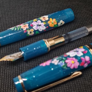 May include: A teal fountain pen featuring a floral pattern. The pen's body and cap are decorated with hand-painted flowers in pink, orange, and white. It has a gold nib and a transparent ink reservoir.