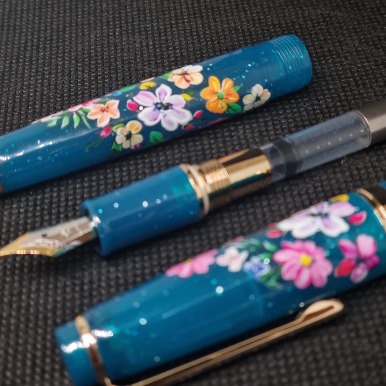 May include: A teal fountain pen featuring a floral pattern. The pen's body and cap are decorated with hand-painted flowers in pink, orange, and white. It has a gold nib and a transparent ink reservoir.