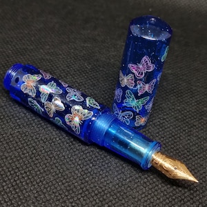 Puede incluir: Una pluma estilogr&aacute;fica azul con plum&iacute;n dorado y capuch&oacute;n. El bol&iacute;grafo est&aacute; decorado con dise&ntilde;os de mariposas iridiscentes. El cuerpo del bol&iacute;grafo es azul transl&uacute;cido y el capuch&oacute;n est&aacute; quitado, revelando el plum&iacute;n.