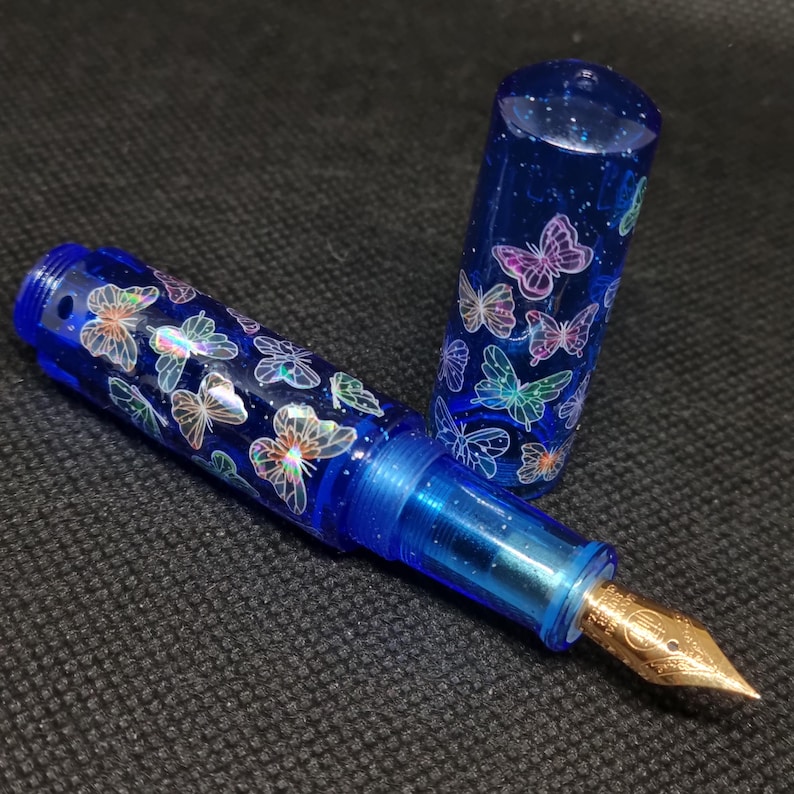 Puede incluir: Una pluma estilogr&aacute;fica azul con plum&iacute;n dorado y capuch&oacute;n. El bol&iacute;grafo est&aacute; decorado con dise&ntilde;os de mariposas iridiscentes. El cuerpo del bol&iacute;grafo es azul transl&uacute;cido y el capuch&oacute;n est&aacute; quitado, revelando el plum&iacute;n.