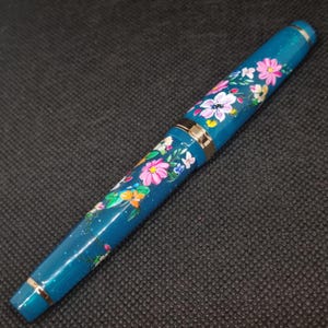 May include: A teal fountain pen with a floral pattern. The pen has gold-coloured bands and accents. The flowers are painted in pink, white, yellow, and orange. The pen has a glossy finish and a classic design.