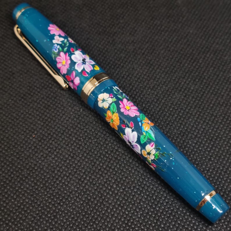 May include: A teal fountain pen with a floral design. The pen has a gold clip and accents, and is decorated with hand-painted flowers in shades of pink, white, orange, and yellow. The pen has a smooth, glossy finish.