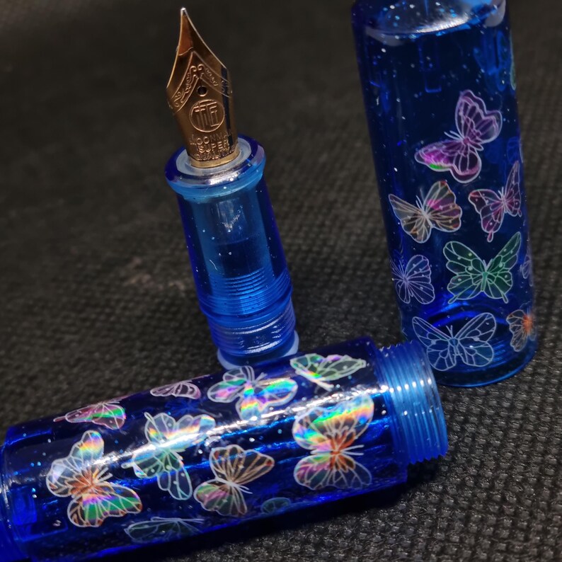 Puede incluir: Una pluma estilogr&aacute;fica azul con plum&iacute;n dorado. El cuerpo y el capuch&oacute;n de la pluma est&aacute;n decorados con dise&ntilde;os de mariposas iridiscentes. La pluma est&aacute; desmontada, mostrando el plum&iacute;n, el cuerpo y el capuch&oacute;n por separado. Es un instrumento de escritura.