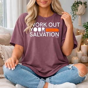 May include: A maroon t-shirt with the text "WORK OUT YOUR SALVATION" in white. The text is above a small orange rectangle with the text "Philippians 2:12". The shirt is paired with blue jeans.