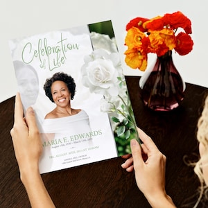 May include: A memorial program titled "Celebration of Life" held by a person. The program features a photo of a smiling woman, white roses, and the name "Maria E. Edwards." A vase of orange flowers sits nearby.