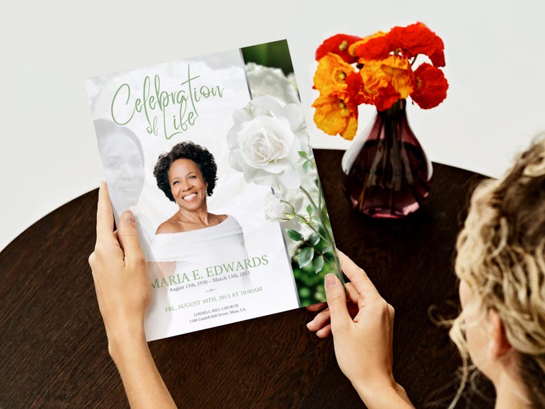 May include: A memorial program titled "Celebration of Life" held by a person. The program features a photo of a smiling woman, white roses, and the name "Maria E. Edwards." A vase of orange flowers sits nearby.
