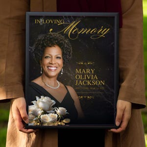 Puede incluir: Una placa conmemorativa enmarcada en negro con una inscripci&oacute;n dorada que dice "In Loving Memory Mary Olivia Jackson" y una foto de una mujer con un ramo de rosas blancas.