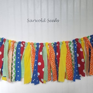 May include: A colorful fabric banner with red, yellow, blue, green, and orange fabric strips. The banner has different patterns, including polka dots, stripes, and chevron. The text "Sarwold Seeds" is printed on a white background above the banner.