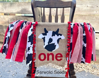 Könnte beinhalten: Ein Banner aus Sackleinen mit einer Kuh-Silhouette und dem Wort "one" in Rot. Das Banner ist mit rot-weißem Gingham-Band, schwarz-weißem Kuhmusterband und roten Pompons verziert. Das Banner hängt an einem Holzstuhl. Der Text "Sarwold Seeds" ist auf das Banner gedruckt.