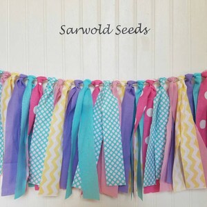 May include: A colorful fabric banner with pink, yellow, purple, and blue fabric strips with different patterns, including polka dots, zigzags, and stripes. The banner is strung on a white background and says "Sarwold Seeds" at the top.
