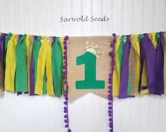 May include: A burlap banner featuring a green number one and a gold glitter crown. The banner is adorned with purple, green, gold, and yellow fabric streamers.