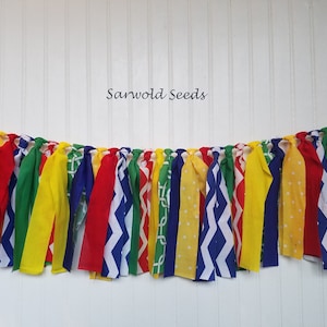 May include: A colorful fabric banner with red, yellow, blue, green, and white fabric strips. The fabric strips have different patterns, including polka dots, zigzags, and geometric designs. The banner is perfect for a party or celebration.