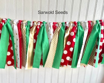 May include: A festive Christmas banner with red, green, gold, and white fabric strips. The banner is made of various patterns including polka dots, chevron, and solid colors. The banner reads "Sarwold Seeds".