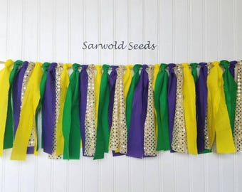 May include: A Mardi Gras party banner with yellow, purple, and green fabric strips and gold glitter fabric strips. The banner says "Sanwold Seeds" at the top.