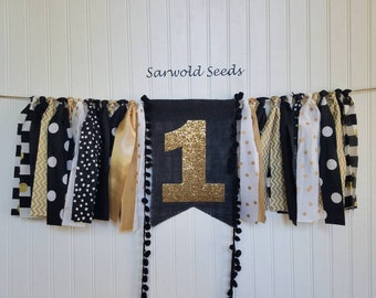 Peut inclure: Une bannière noire et dorée avec le chiffre "1" en paillettes dorées, entourée de bandes de tissu noires et dorées à pois, rayures et chevrons. La bannière est pour une fête de premier anniversaire.