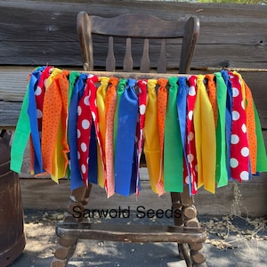 May include: A colorful high chair banner with alternating fabric strips in red polka dots, orange, yellow, green, and blue. The banner is attached to a wooden high chair, perfect for a birthday party or celebration.