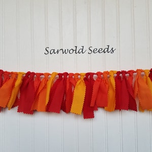 Puede incluir: Una guirnalda de tela con tiras de tela roja, naranja y amarilla atadas juntas. La guirnalda est&aacute; colgada en una pared blanca con el texto "Sarwold Seeds" encima.