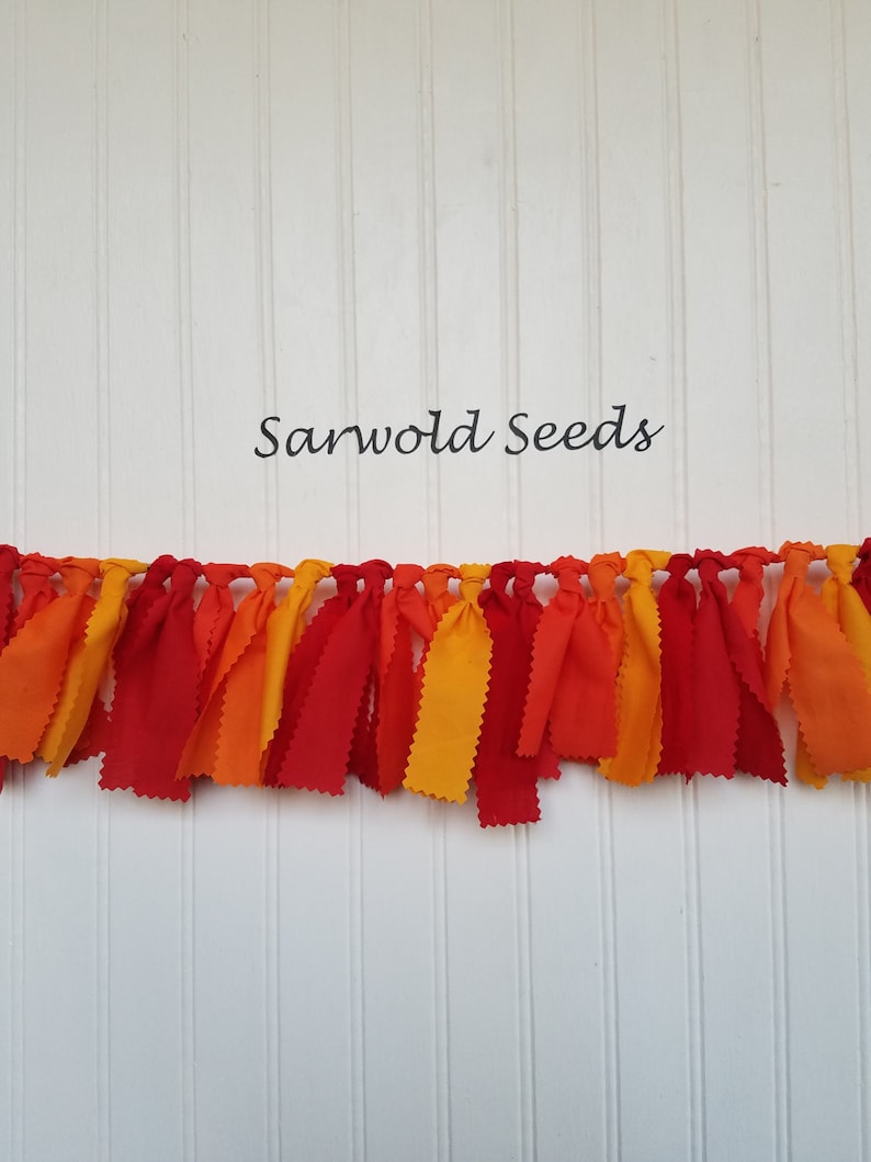 Puede incluir: Una guirnalda de tela con tiras de tela roja, naranja y amarilla atadas juntas. La guirnalda est&aacute; colgada en una pared blanca con el texto "Sarwold Seeds" encima.