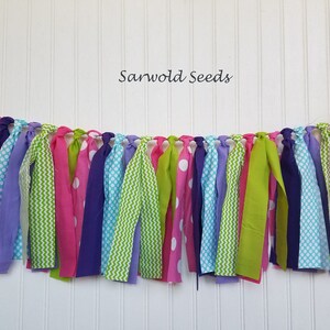 May include: A colorful fabric banner with green, pink, purple, and blue fabric strips with different patterns, including polka dots, zigzags, and stripes. The banner is strung together with white string and has the text "Sarwold Seeds" at the top.