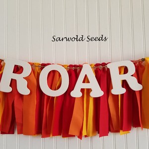 以下が含まれることがあります： 「ROAR」という文字が大きく書かれた白い木製の横断幕。赤い、オレンジ色の、黄色の布のストリップで飾られています。