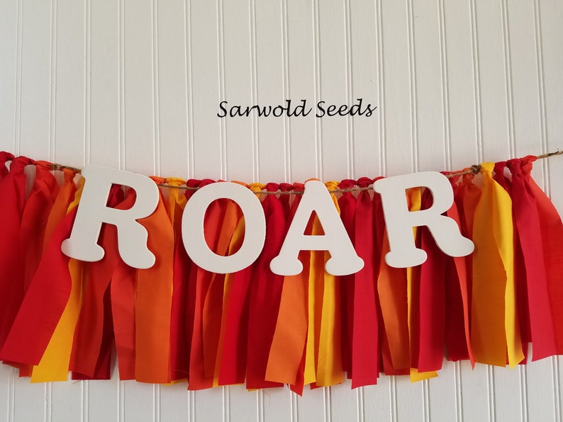 以下が含まれることがあります： 「ROAR」という文字が大きく書かれた白い木製の横断幕。赤い、オレンジ色の、黄色の布のストリップで飾られています。