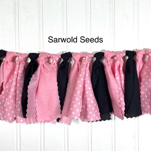 Puede incluir: Una pancarta de tela rosa y negra con lunares blancos. La pancarta est&aacute; atada con nudos y tiene un borde festoneado. El texto "Sarwold Seeds" est&aacute; impreso sobre un fondo blanco por encima de la pancarta.