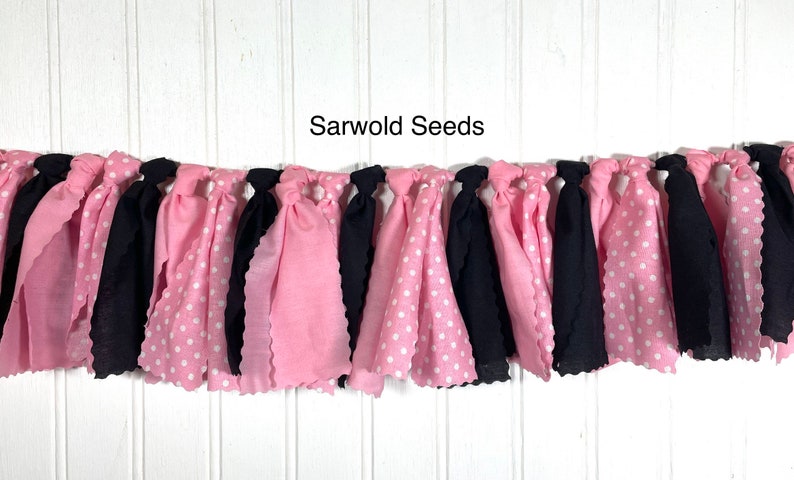 Puede incluir: Una pancarta de tela rosa y negra con lunares blancos. La pancarta est&aacute; atada con nudos y tiene un borde festoneado. El texto "Sarwold Seeds" est&aacute; impreso sobre un fondo blanco por encima de la pancarta.