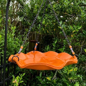 May include: An orange, wavy-edged bird feeder suspended by black chains. The feeder is decorated with colorful beads and hangs in front of green foliage. The feeder is designed to attract birds to a garden.
