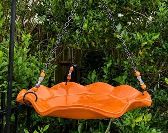 Könnte beinhalten: Eine orangefarbene Vogelfutterstelle mit gewelltem Rand, die an schwarzen Ketten aufgehängt ist. Die Futterstelle ist mit bunten Perlen verziert und hängt vor grünem Laub. Sie soll Vögel in einen Garten locken.