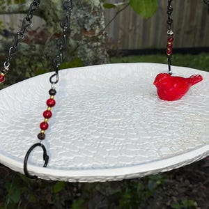 May include: A white, textured bird bath with a red bird figurine in the center. The bird bath is suspended by black chains with decorative red and gold beads. The bird bath is hanging from a tree branch.