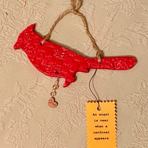 May include: Red cardinal bird ornament with a heart-shaped tag that reads "An angel is near when a cardinal appears". The ornament is made of clay and has a textured surface. It is hanging from a twine string.