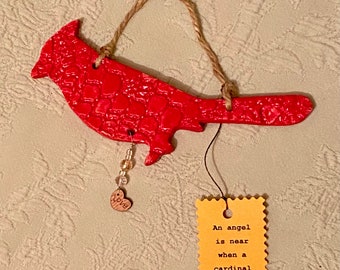 May include: A red cardinal-shaped ornament with a textured surface, hanging from a twine loop. A small tag with the text "An angel is near when a cardinal appears" is attached. A beaded charm with a heart that says "love" is also attached.