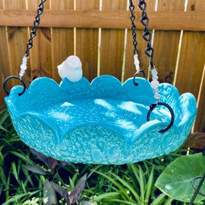 May include: A turquoise blue ceramic bird bath with a scalloped edge hangs from a black chain with white beads. The bird bath is filled with water and has a white ceramic bird perched on the edge.