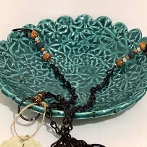 May include: A teal ceramic hanging planter with a scalloped edge and intricate floral design. The planter has two black chains with brown wooden beads attached to it.