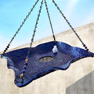 May include: A hanging blue bird feeder with a textured surface and a small white bird figurine. The feeder is suspended by black chains and adorned with decorative beads. The feeder has a diameter of approximately 12 inches.