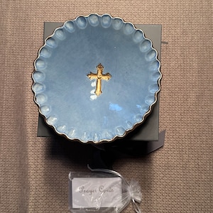 Puede incluir: Un plato de cerámica azul con un borde dorado y una cruz dorada en el centro. El plato está en una caja negra con una cinta negra. Una pequeña tarjeta blanca con el texto "Prayer Cards" está en una bolsa de plástico transparente.