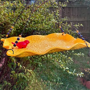 May include: A bright yellow, textured bird feeder with a wavy edge, suspended by black chains. A small, red bird figurine sits on the feeder. The chains are adorned with decorative beads in shades of blue, gold, and brown. The feeder hangs in front of green foliage.