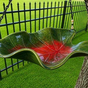 May include: A ceramic hanging bird bath shaped like a large leaf with a glossy green exterior and a vibrant red center. The basin features raised veins and is suspended by black metal chains adorned with small beige, clear, and green beads.