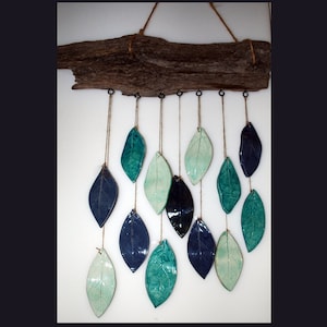 May include: A driftwood wall hanging with ceramic leaf ornaments in shades of teal, blue, and green. The leaves are suspended from the driftwood by twine.